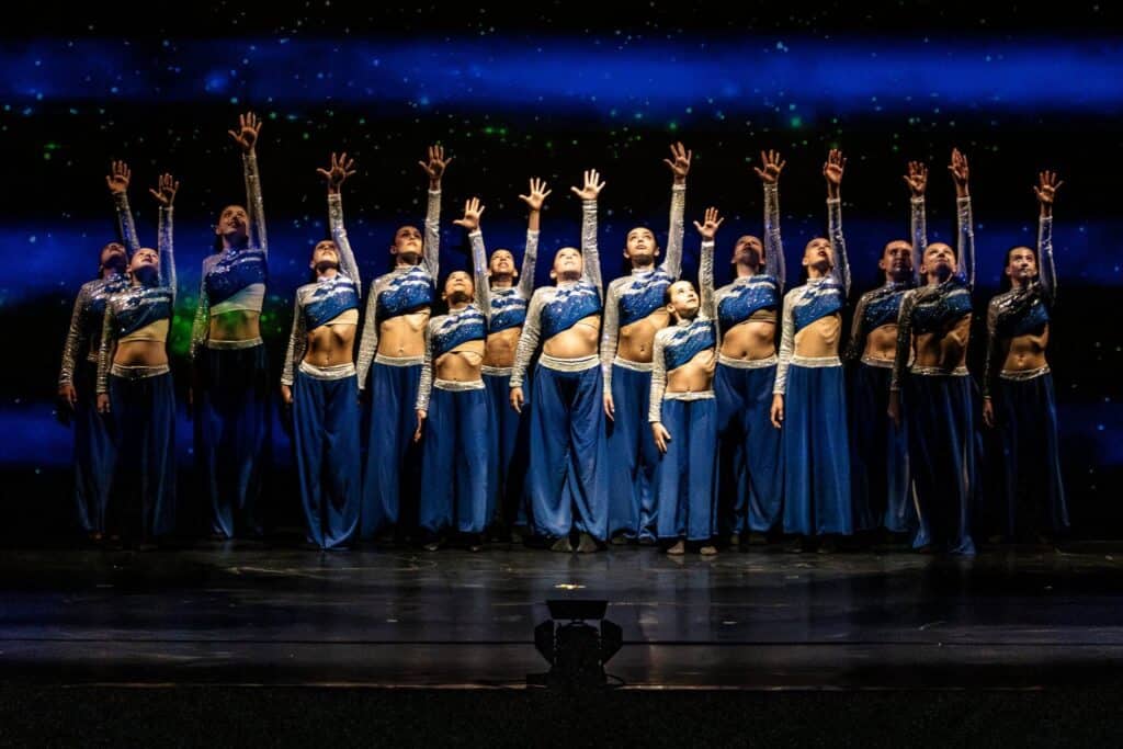 Dancer in light blue top leads energetic movement with two performers behind her in moody purple lighting.