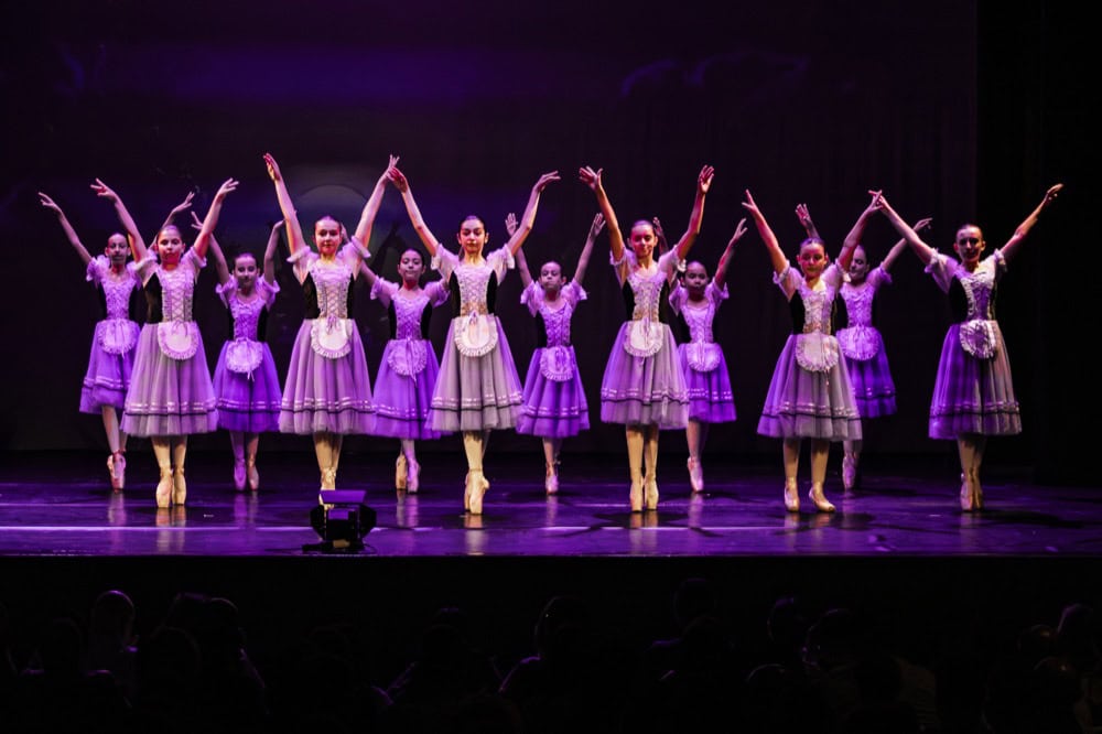Ballet dancer in white tutu performs en pointe center stage with ensemble in blue lighting and stage lights.