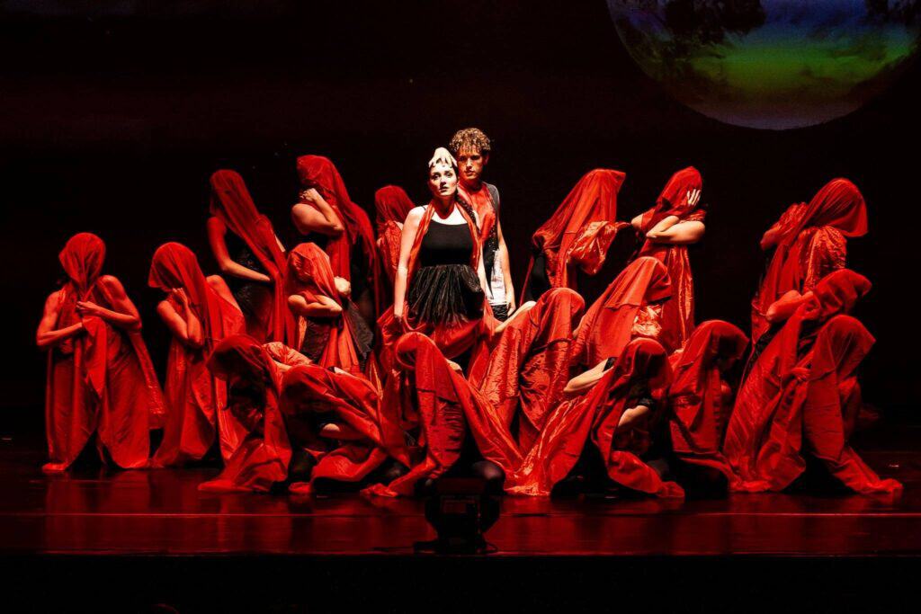 Ballet dancers in white tutus performing with raised arms on a dark stage with audience visible