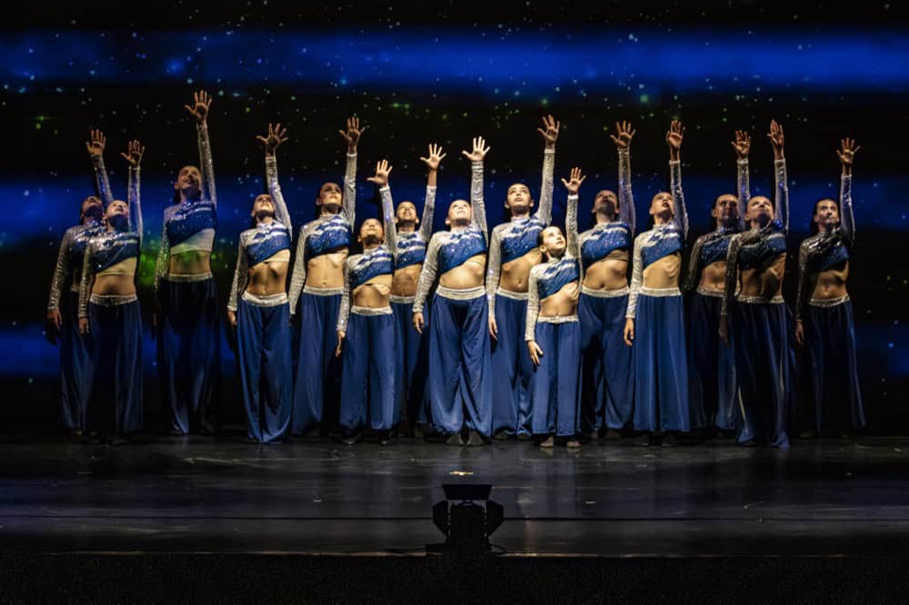 Dancer in light blue top leads energetic movement with two performers behind her in moody purple lighting.