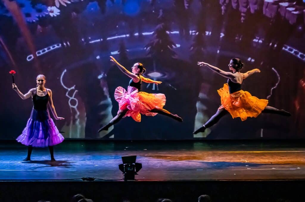 Ballet dancers in white tutus performing with raised arms on a dark stage with audience visible